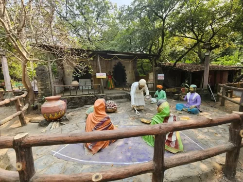 Sai Baba serving food to devotees statue scene at Sai Heritage Village near Naini Lake Nainital, featuring women in colorful saris around traditional hut, perfect cultural heritage experience Uttarakhand tour package.