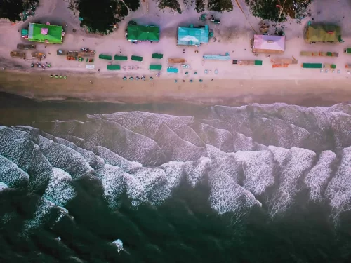 Drone aerial view of Mandrem Beach North Goa during golden hour sunset, featuring colorful shacks beach umbrellas chairs palm trees white sandy shore Arabian Sea waves, perfect serene beach shack experience with Mandrem Beach Goa tour package.
