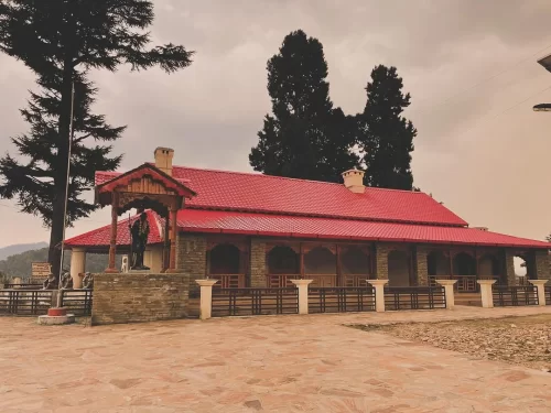 Anasakti Ashram Kausani stone red roof building Gandhi statue wooden pavilion deodar trees cloudy skies distant hills, perfect Gandhi spiritual retreat, Uttarakhand tour packages.