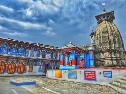 Ukhimath Omkareshwar Temple towering stone shikhara colorful blue red yellow wood carved facade cloudy skies flag fluttering, perfect Kedarnath winter Shiva temple, Uttarakhand tour packages.