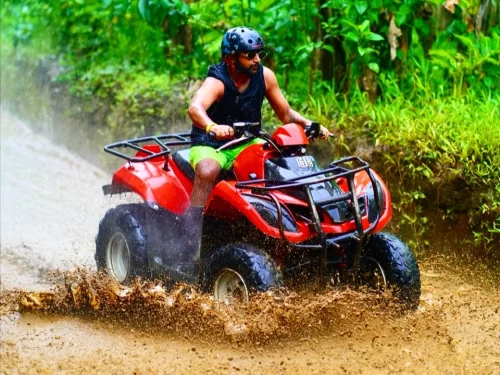 Man riding a red ATV through muddy forest trail, splashing water, wearing helmet and sunglasses during an adventurous off-road ride.