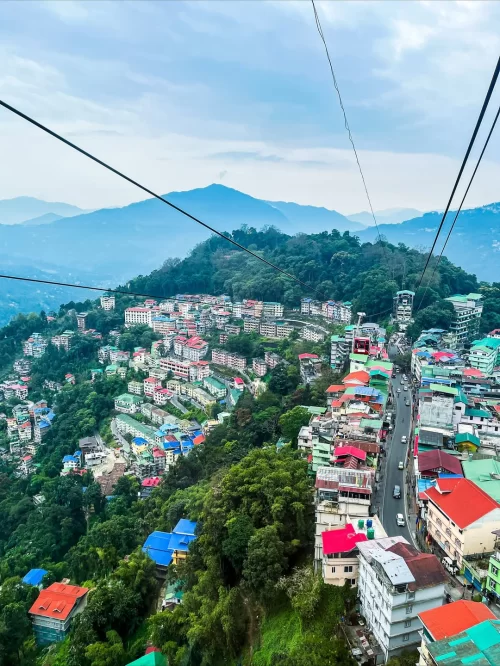 Aerial view of Gangtok city with colorful hillside buildings and cable car lines against misty mountains, a vibrant capital experience featured in Sikkim tour packages.