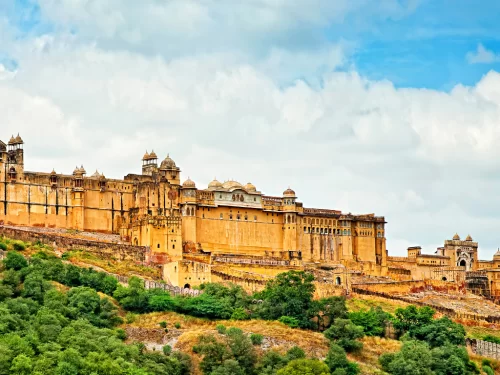 Full facade view of Amber Fort (Amer Fort) in Jaipur Rajasthan on Aravalli hillside with multi-tiered yellow sandstone palaces, towers amid greenery under partly cloudy blue sky, perfect Rajasthan tour package.