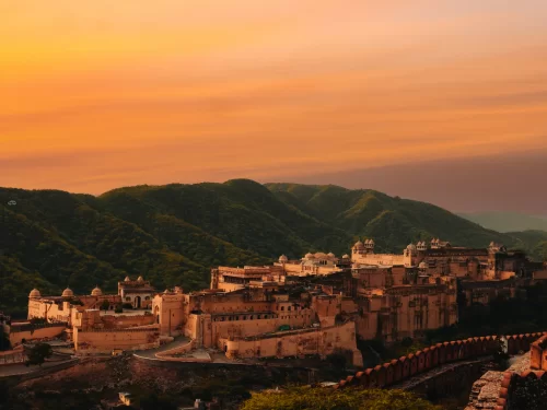 Sunset panoramic view of Nahargarh Fort in Jaipur Rajasthan atop Aravalli hills with pinkish sandstone structures, walls amid green slopes under orange purple sky, perfect Rajasthan tour package
