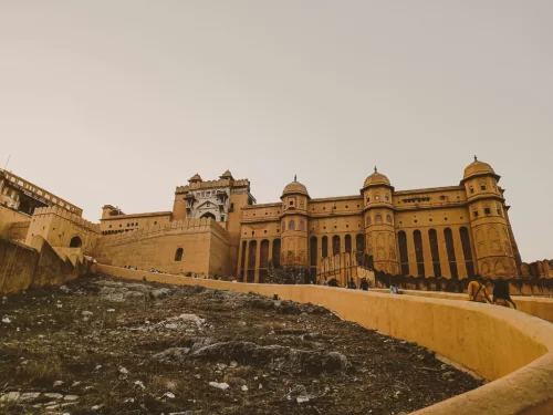 Golden hour view of Amer Fort (Amber Fort) in Jaipur Rajasthan showcasing majestic sandstone facade, domes, ramparts and hillside path with visitors under hazy sky, perfect Rajasthan tour package