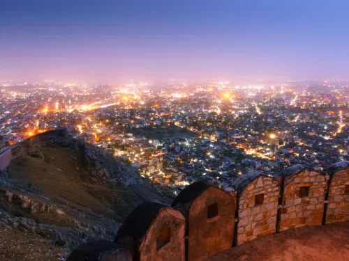Night panoramic view from Nahargarh Fort overlooking twinkling Jaipur city lights, Aravalli hills and fort walls under twilight purple sky, perfect Rajasthan tour package.