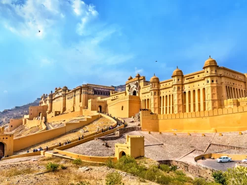 Daytime panoramic view of Amber Fort (Amer Fort) in Jaipur Rajasthan with expansive yellow sandstone walls, gates, towers on hillside amid hills and blue sky with clouds, perfect Rajasthan tour package.