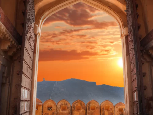 Sunset through arched gateway at Amber Fort in Jaipur Rajasthan framing orange sky, setting sun and distant hill fort silhouette with ramparts foreground, perfect Rajasthan tour package.