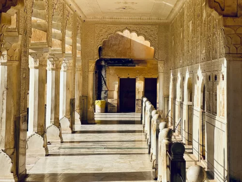 Golden sunlight illuminating ornate marble colonnade corridor with intricate arches and pillars at Amber Fort in Jaipur Rajasthan leading to shadowed doorway, perfect Rajasthan tour package.