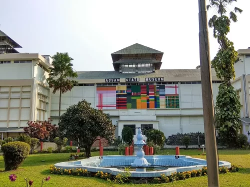 Assam State Museum main building at Guwahati during sunny day, featuring colorful mural fountain palm trees, perfect cultural experience Assam tour package. 
