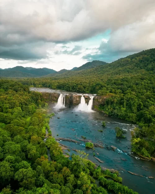 Aerial view of Athirappilly Waterfalls in Kerala, with twin cascades plunging into a wide river surrounded by dense green forest and rolling hills under a cloudy sky.
