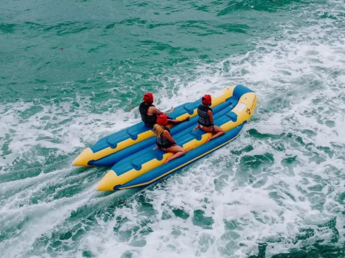 Three people wearing life jackets and helmets enjoying a banana boat ride over turquoise ocean waves, being pulled at speed across the sea.