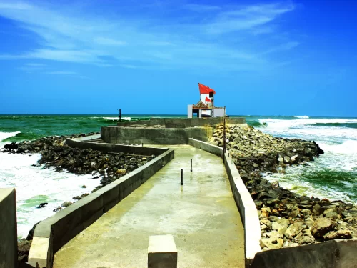 Bhadkeshwar Mahadev Temple Dwarka concrete pathway leading to white shrine with red saffron flags amid crashing Arabian Sea waves on rocky breakwater under clear blue skies, unique ocean Shiva temple perfect for Gujarat tour packages.