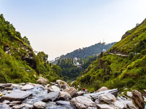 Trekkers on Bhagsu Nag Waterfall pathway at Dharamshala during golden hour, featuring lush green valleys, rocky stream, hills, perfect adventure Himachal Pradesh tour package.