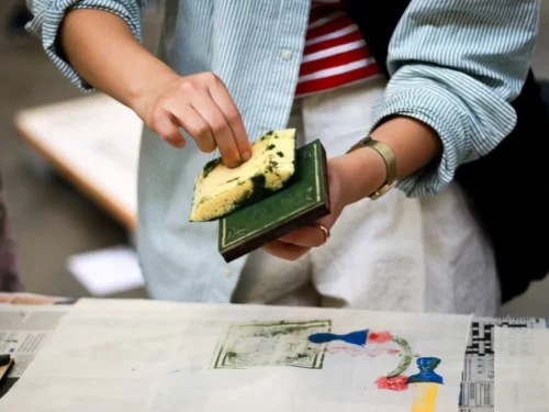 A person participating in a block printing workshop, applying ink to a carved wooden block and stamping colorful patterns onto fabric.