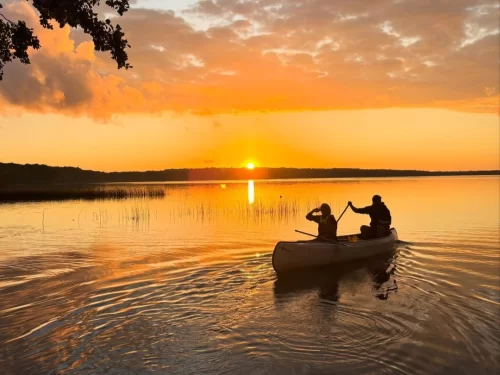 Canoeing couple paddling a canoe on calm lake waters during golden sunset with scenic sky reflections and peaceful surroundings.