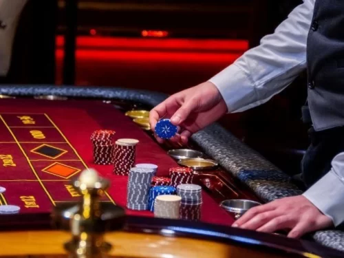 A casino scene showing a dealer placing chips on a red felt gaming table, with neatly stacked poker chips and betting areas under warm ambient lighting.