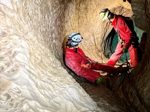 Caving adventure with two explorers in protective helmets and red gear rappelling inside a narrow limestone cave tunnel with rugged rock formations.