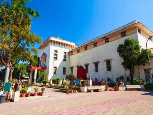 Central Museum Indore Madhya Pradesh in sunny weather, featuring white building red entrance, trees statues gardens, perfect cultural Madhya Pradesh tour package.