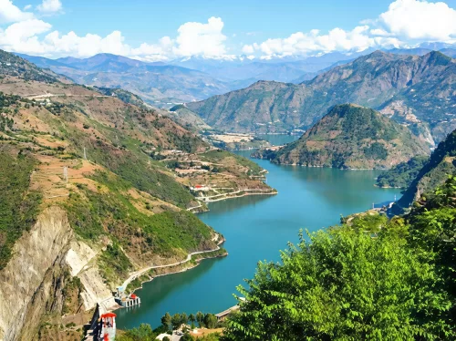 Chamera Lake in Dalhousie featuring a winding turquoise reservoir surrounded by rolling hills, mountain roads, and scenic Himalayan ranges under a bright blue sky.