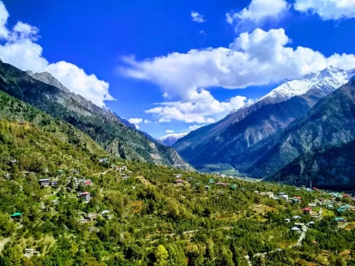 Panoramic view from Chitkul View Point showcasing lush green valley slopes dotted with houses, winding mountain roads, and snow-capped Himalayan peaks under a bright blue sky with scattered clouds.