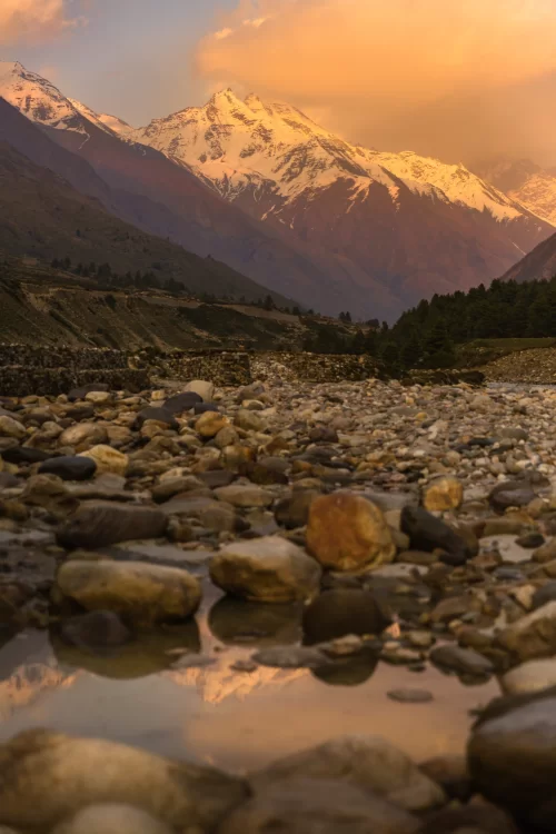 Snow capped Himalayan peaks glowing at sunset above rocky riverbed in Chitkul, a serene mountain village featured in Himachal Pradesh tour packages.