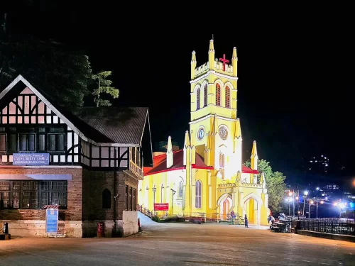 Christ Church Shimla Neo-Gothic yellow church tower illuminated at night with red cross adjacent colonial building trees city lights, perfect evening heritage view, Himachal Pradesh tour packages.