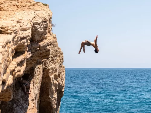 Cliff Jumping thrill as a man leaps backward off a rocky seaside cliff into the blue ocean under a clear sky.