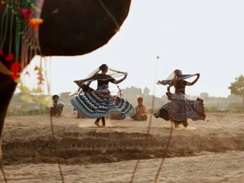 Cultural Folk Dance performance with women in vibrant traditional attire twirling gracefully in a desert village setting at sunset.