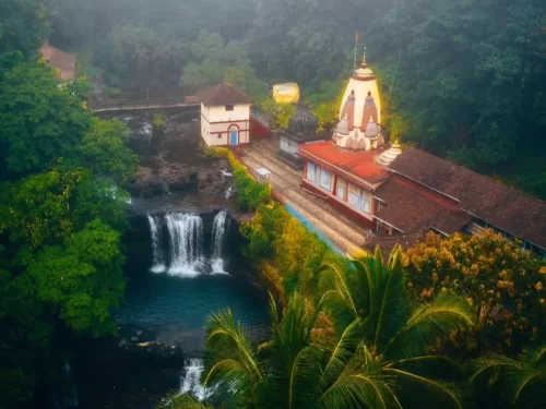 Dhootapapeshwar Mandir in Maharashtra surrounded by lush greenery and a scenic waterfall beside the temple complex, a serene spiritual spot featured in Maharashtra tour packages 
