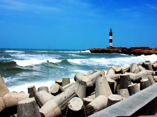 Dwarka Lighthouse on Arabian Sea coast during clear day, featuring black-white striped tower amid crashing waves and breakwater blocks, perfect coastal experience with Gujarat tour packages.