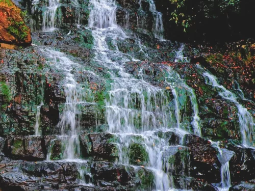 Elephant Falls Shillong Meghalaya tiered waterfall cascading over mossy green rocks orange toned water flow dark stone ledges forested backdrop vibrant natural beauty, perfect Meghalaya tour packages.