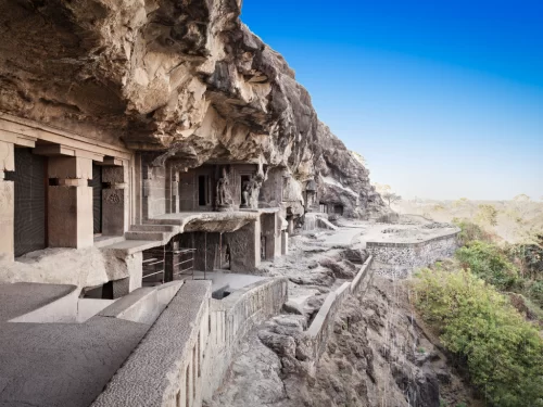 Jain caves series carved into cliffside at Ellora Caves during clear blue sky, featuring rock facades, wall, greenery, perfect UNESCO Aurangabad tour package. 