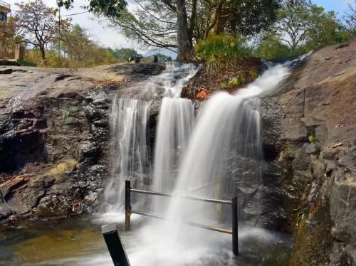 Fairy Falls Kodaikanal Pambar River multi-tier cascading waterfall rocky cliffs lush green trees metal railing pool base scenic picnic spot Tamil Nadu hill station tour packages.