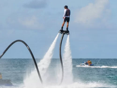 A person flyboarding over the ocean, propelled high above the water by powerful water jets from a connected hose, with a jet ski in the background on a clear day.