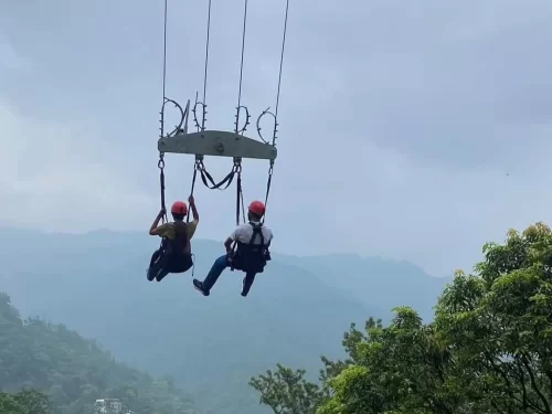 Giant Swing two adventure seekers suspended midair on a high swing ride overlooking misty green hills and forest landscape.