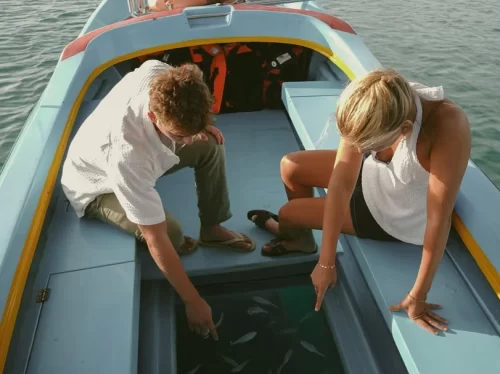 Glass Bottom Boat ride couple looking through transparent boat floor at fish swimming in clear blue sea water.