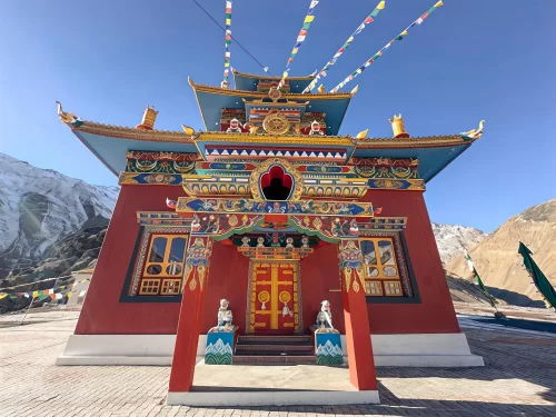 Gue Monastery Kaza Spiti close-up winter, featuring vibrantly painted maroon temple golden roofs prayer flags snowy terrain distant mountains blue skies, perfect Himachal Pradesh tour packages.