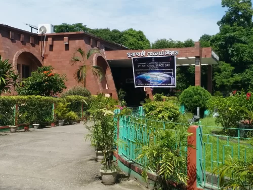 Guwahati Planetarium Assam campus during sunny daytime, featuring brick building, green gardens and National Space Day banner, perfect family experience Guwahati tour package. 
