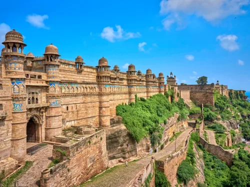 Gwalior Fort ramparts during clear afternoon, featuring turquoise tilework pavilions and domes atop cliffside with lush greenery, perfect heritage experience in Madhya Pradesh tour package.