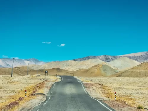 Hanle Ladakh highway barren desert mountains blue sky, winding asphalt road colorful poles dramatic peaks Changthang plains, perfect Hanle observatory road trip package.