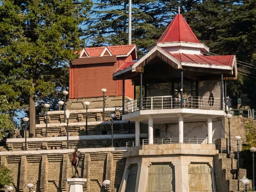 Hawa Ghar in Shangarh featuring a pavilion-style structure with a red sloping roof, stone terraces, surrounding deodar trees, and a statue in the foreground set against a hillside backdrop.