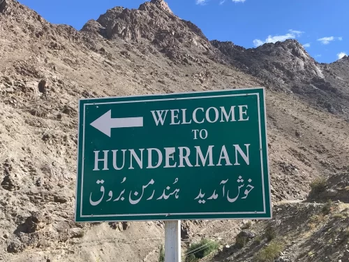 Hunderman Village Kargil Ladakh official green welcome signboard with left arrow in English-Urdu, dramatic barren Himalayan peaks under clear blue skies, LoC ghost village Brokpa settlement entry, perfect adventure India tour package.