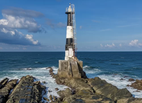 Black white lighthouse on rocky jetty at Ross Island Andaman during partly cloudy day, featuring crashing waves blue ocean, weathered structure, perfect colonial heritage Andaman tour package.
