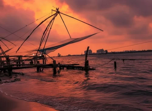 Silhouette Chinese fishing nets at Thekkady during fiery orange sunset, featuring wooden jetty water reflections and clouds, perfect cultural experience Thekkady tour package.