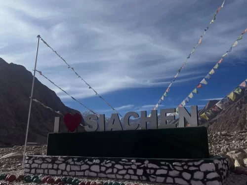 I Love Siachen large signboard red heart white letters prayer flags fluttering poles Karakoram mountains cloudy sky rocky terrain tourist photo spot Siachen Base Camp Ladakh Nubra Valley. (162 chars) ​