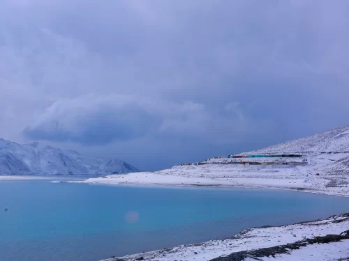 Pangong Tso Lake Ladakh winter snowy landscape turquoise water blue ice shore snow covered mountains terraced hills multi color train blue purple cloudy sky, high altitude frozen lake adventure tour package.