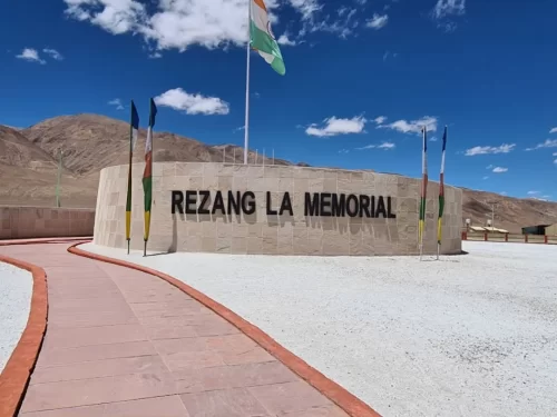 Rezang La War Memorial at Rezang La Pass Ladakh during clear skies, featuring Indian Tricolour flag, prayer flags, stone wall, Himalayan peaks, perfect patriotic experience with Ladakh tour package. 