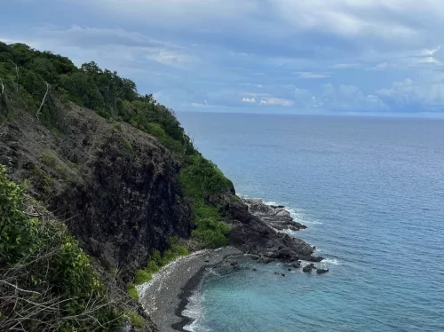 Cliffside view at Chidiya Tapu Port Blair during partly cloudy day, featuring black rocky cliffs, turquoise beach cove, lush green vegetation, ocean waves, perfect nature adventure Andaman tour package.