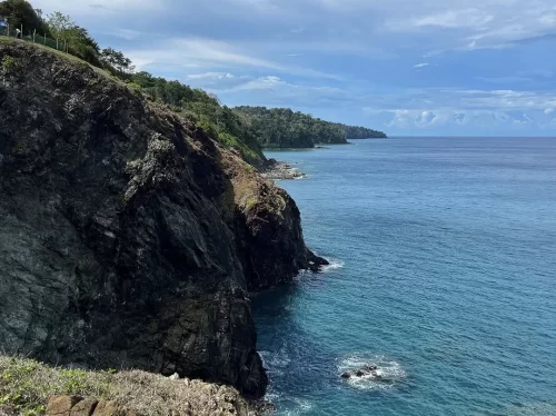 Seaside cliffs at Chidiya Tapu Port Blair during partly cloudy skies, featuring rugged black rocks, turquoise ocean waves, dense green forest, distant beaches, perfect scenic adventure Andaman tour package.